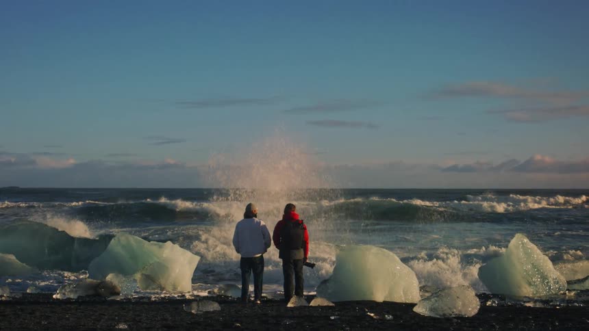 A man with a camera stands with another man and watches waves crash on shore