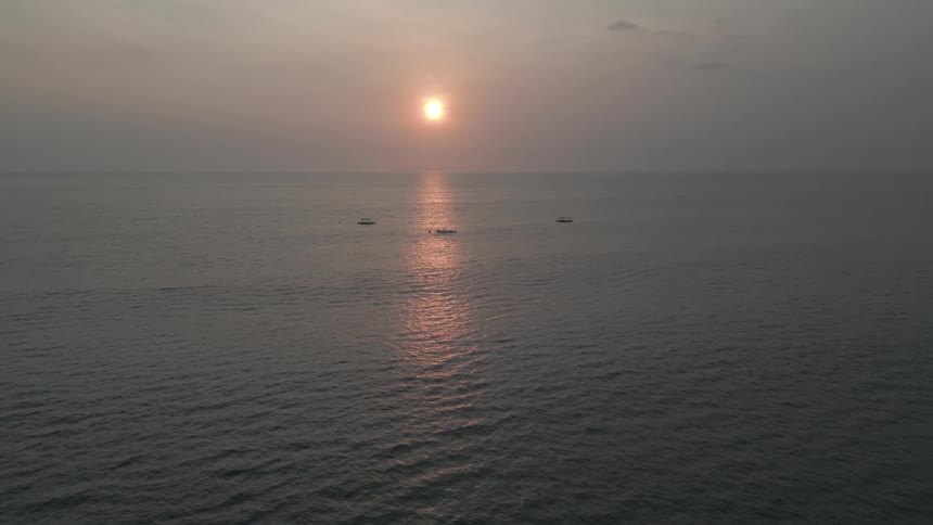 The sun sets over a calm expansive ocean its light reflecting on the waters surface with three small boats visible in the distance