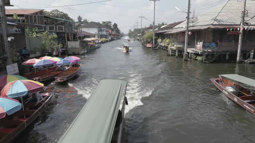 Market life unfolds on Thai waterways, as a longtail boat moves down a narrow canal lined with waterfront homes and colorful market boats as seen from an overhead bridge gradually revealing the expanse of water under a bright tropical sky