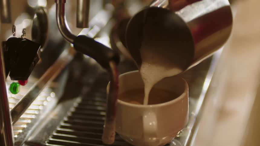 A barista pours steamed milk from a metal pitcher into a coffee cup over an espresso machine