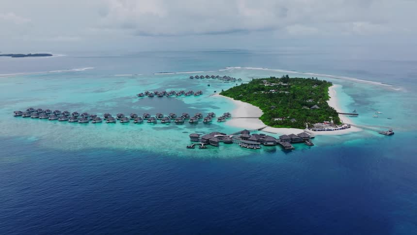 An aerial view shows a lush tropical island surrounded by turquoise shallows and deep blue ocean dotted with clusters of overwater bungalows extending from its shores