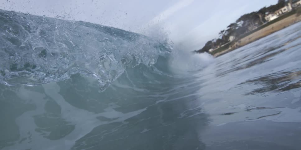 A wave breaking at the shoreline with the waters texture and movement in sharp focus and a blurred cityscape visible in the background