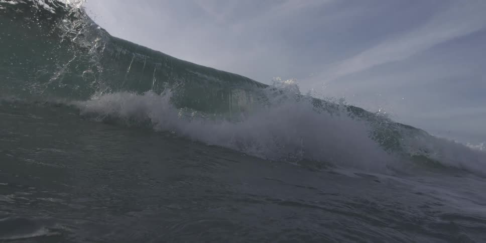 A wave cresting with the dark water reflecting light and a hint of white foam at the top under a gray sky