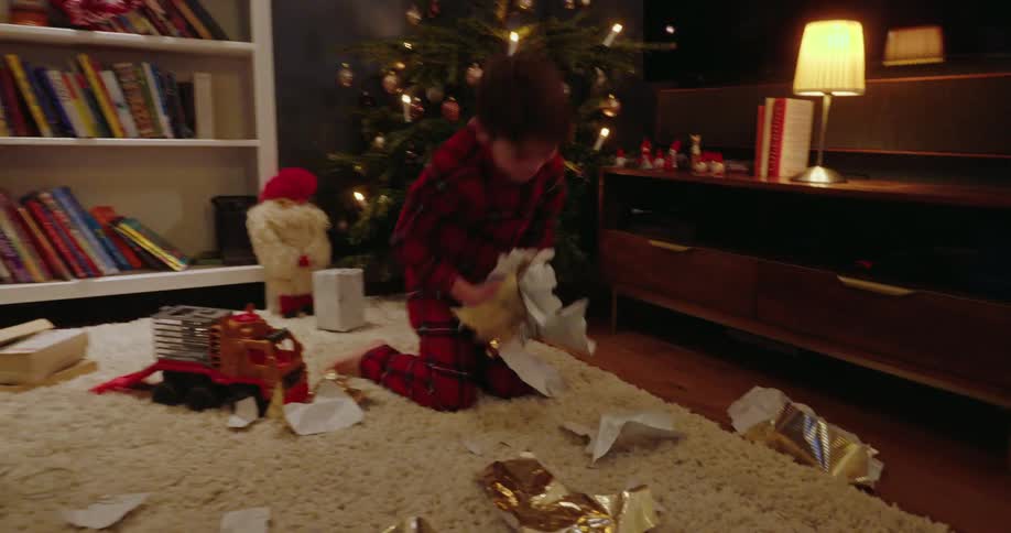 An excited young boy in plaid pajamas throws wrapping paper in the air while opening presents beside a decorated christmas tree