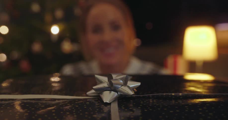 A person receives a christmas gift wrapped in bright silver paper with a bow as a christmas tree stands decorated in the background