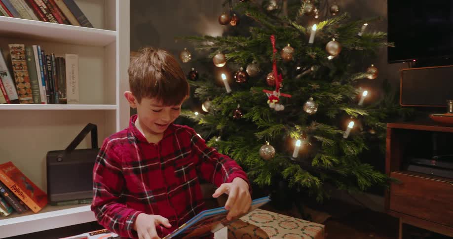 Excited young boy in plaid pajamas unwrapping a book he received as a gift