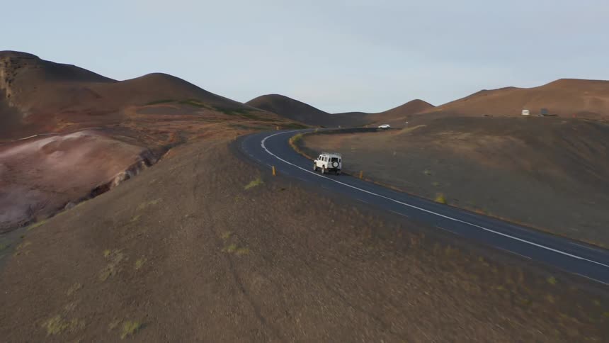 A white jeep drives along a winding road through a vast arid landscape with rolling hills