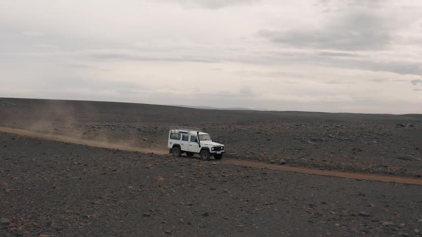 A white land rover defender kicks up dust as it drives along a dirt track through a barren rocky landscape under an overcast sky
