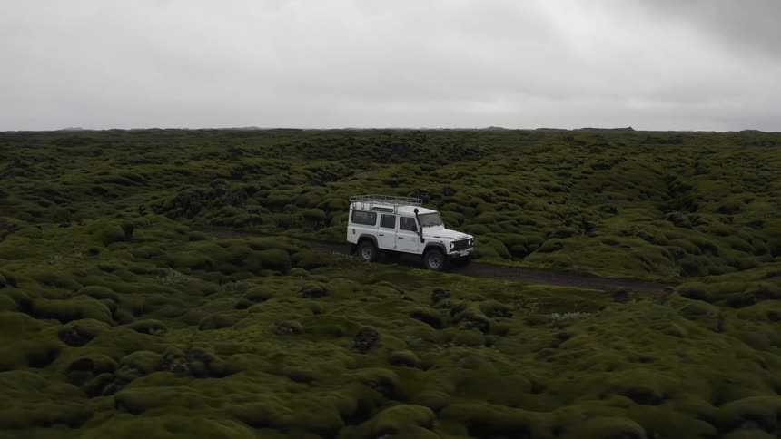 Vehicle moves along a road passing through vegetation covered rough terrain on an overcast day