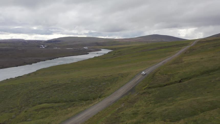 Vehicle travels along a rural highway paralleling the river in hilly terrain on an overcast day