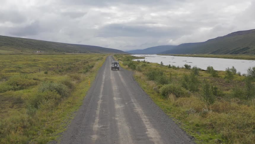 A white land rover defender 90 drives down a gravel road through a vast open landscape with rolling hills and a river in the distance under a cloudy sky