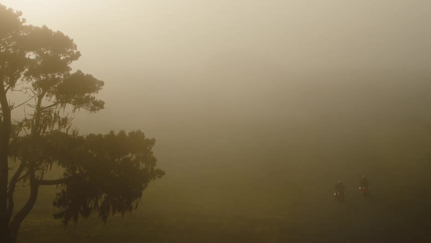 Two motorcycles drive through the very thick fog covering everything