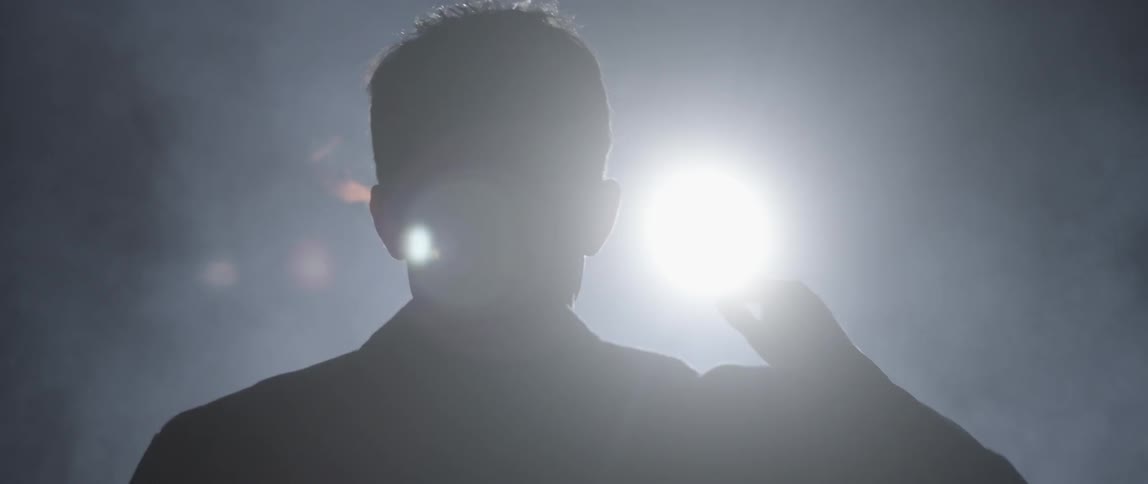 Spectacled man holding a torch in the darkness but then turning around as a bright light approaches him from behind