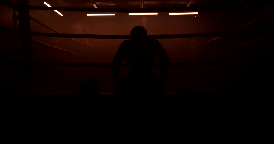 Silhouetted man sitting in a boxing ring takes a deep breath and raises his arms to his head