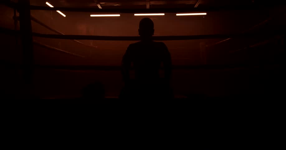 Male boxer silhouetted against flashing lights in a dark gym sits at the edge of the ring and rests