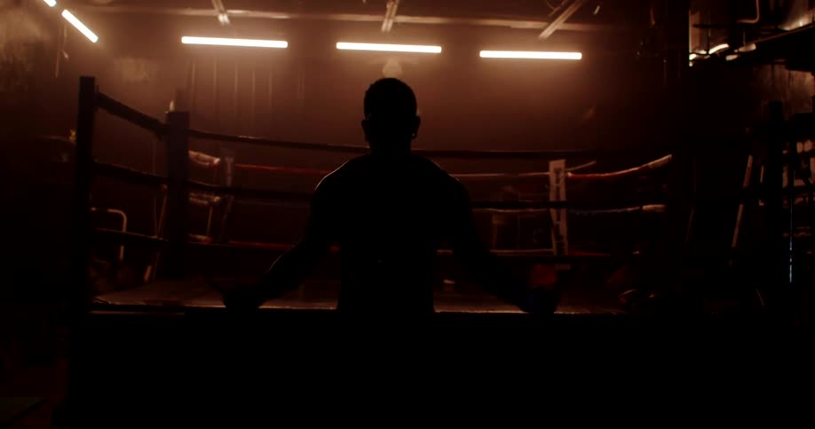 A man is jumping rope in front of a boxing ring in a dark room