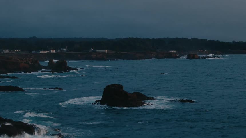 A twilight seascape captures a rocky coastline with a few scattered buildings where dark blue ocean waves meet the shore under a darkening gray sky