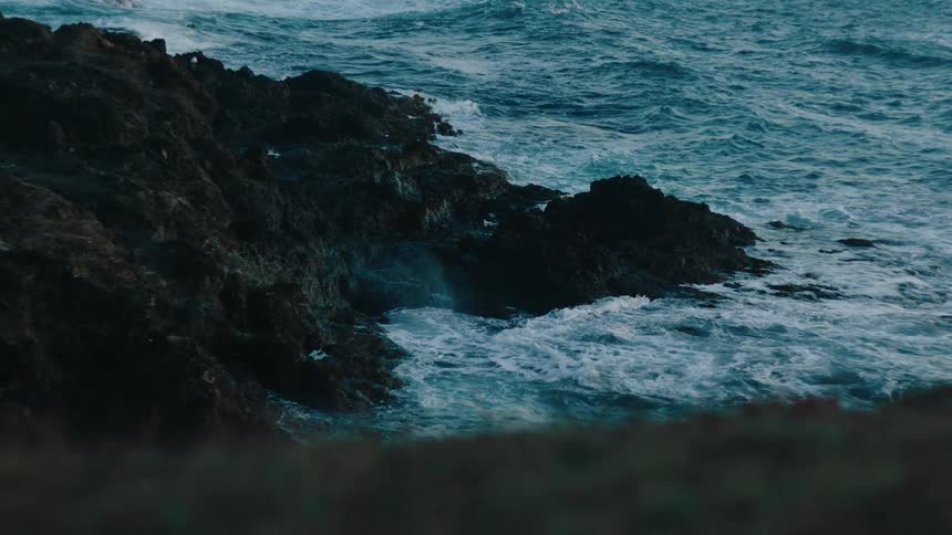 A dark rocky cliff juts into a turbulent blue ocean with white waves crashing against the rocks creating a dynamic contrast between the rugged land and the powerful sea