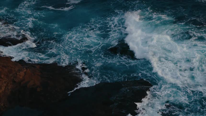 An aerial view of the dramatic contrast of dark jagged rocks against the swirling white foam of crashing waves in the deep blue ocean