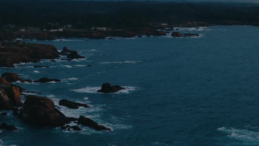 A twilight coastal scene captures dark rocky cliffs plunging into a deep blue ocean with white waves crashing against the rocks under a dimly lit sky