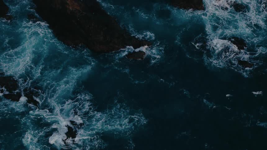 Aerial view of dark jagged rocks surrounded by the swirling white foam of crashing waves against the deep blue ocean creating a dramatic contrast and highlighting the raw power of the sea