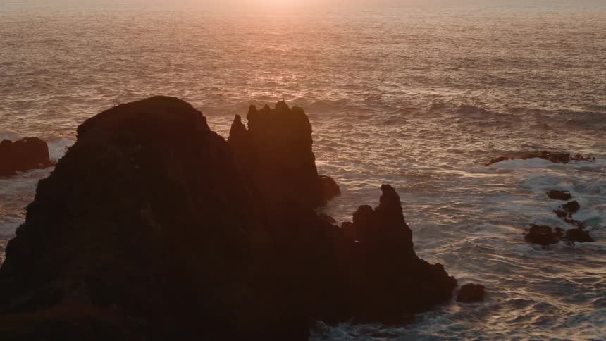 A sunset casts a golden glow over the ocean illuminating jagged rocks emerging from the turbulent water near a dark silhouetted cliff