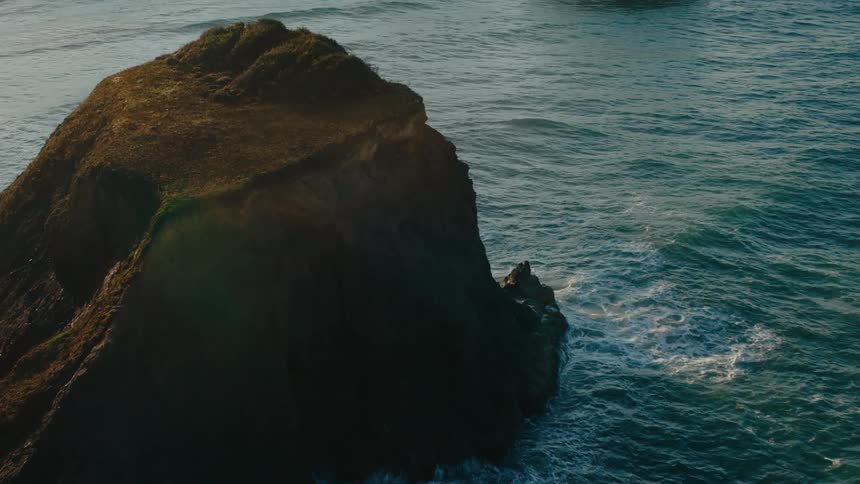 An aerial view of a dark rocky outcropping surrounded by turquoise ocean waves with other rock formations visible in the distance under a hazy sky