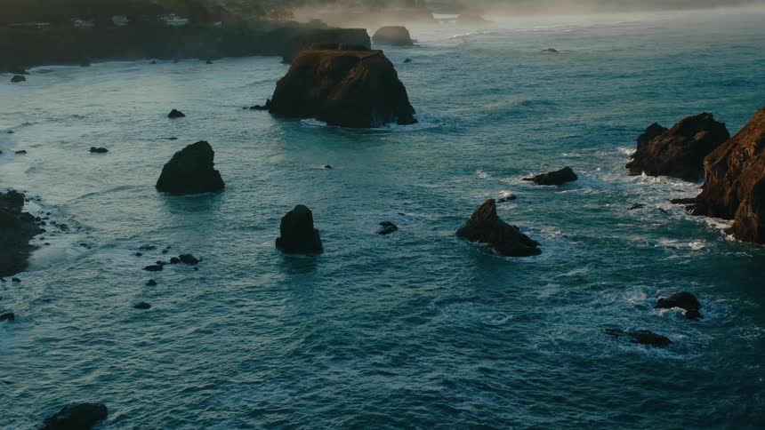 An aerial view of a dramatic coastline with rugged cliffs and rocky outcroppings scattered in the turbulent blue ocean under a hazy sky