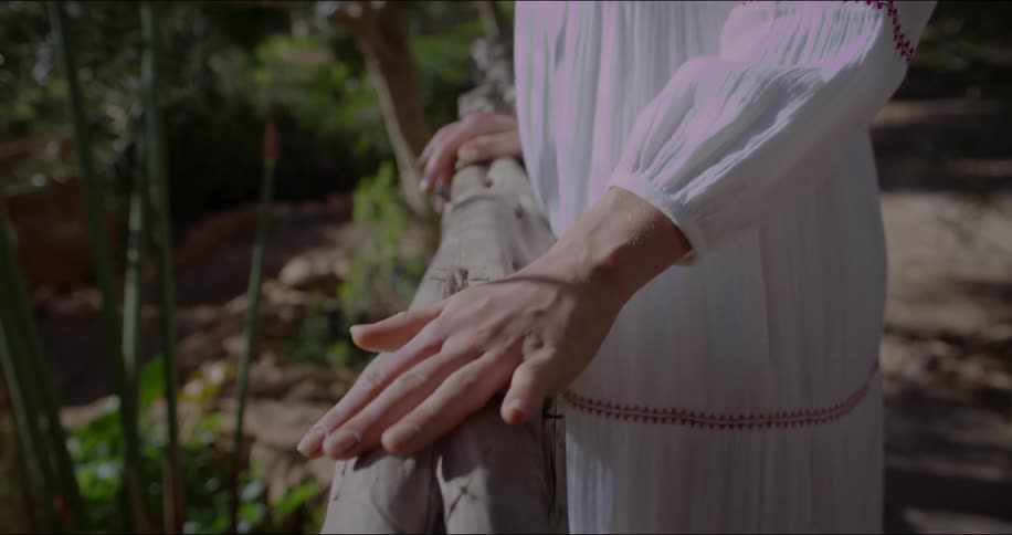 A woman wearing a white dress with red embroidery rests her hand on a wooden railing in an outdoor setting