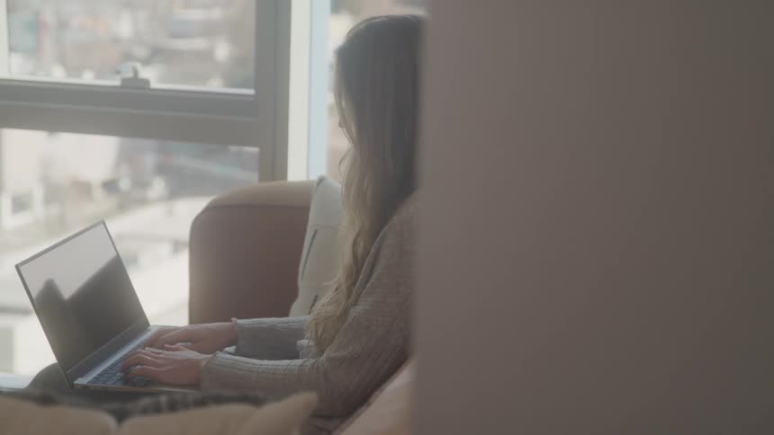 A woman with long hair sits on a couch working on a laptop near a bright window with a blurred foreground creating a sense of intimacy and focus on her activity