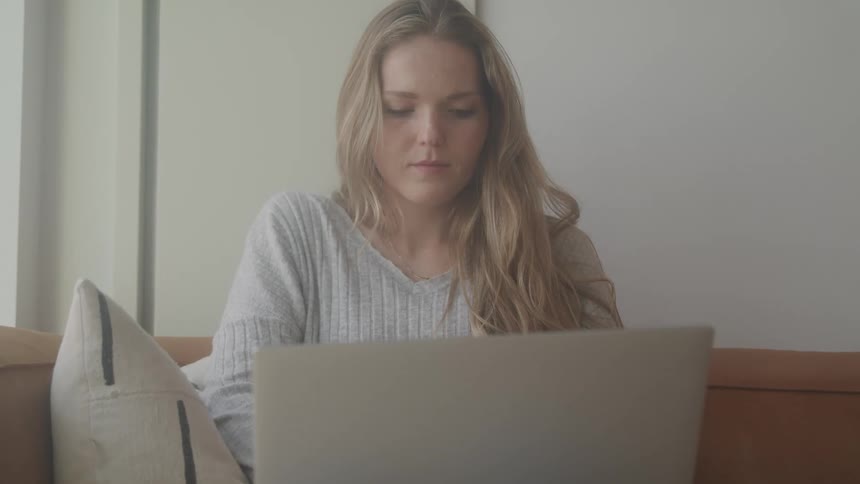 A young woman with long blonde hair wearing a light gray sweater sits on a couch and looks down at a laptop screen concentrating on her work