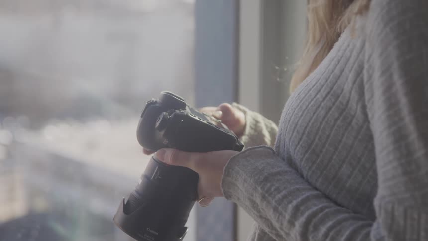 A woman with long blonde hair wearing a gray sweater holds a camera in her hands with a blurred window in the background