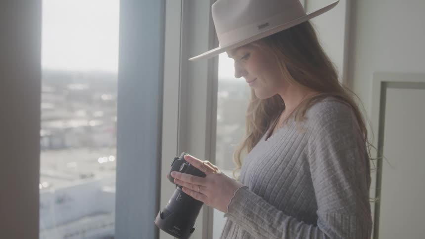 A woman wearing a hat and a gray sweater examines a camera lens while standing near a window overlooking a cityscape