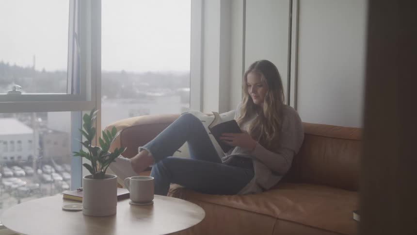 A woman with long blonde hair sits comfortably on a couch by a large window reading a book in a bright modern room with a view of a cityscape