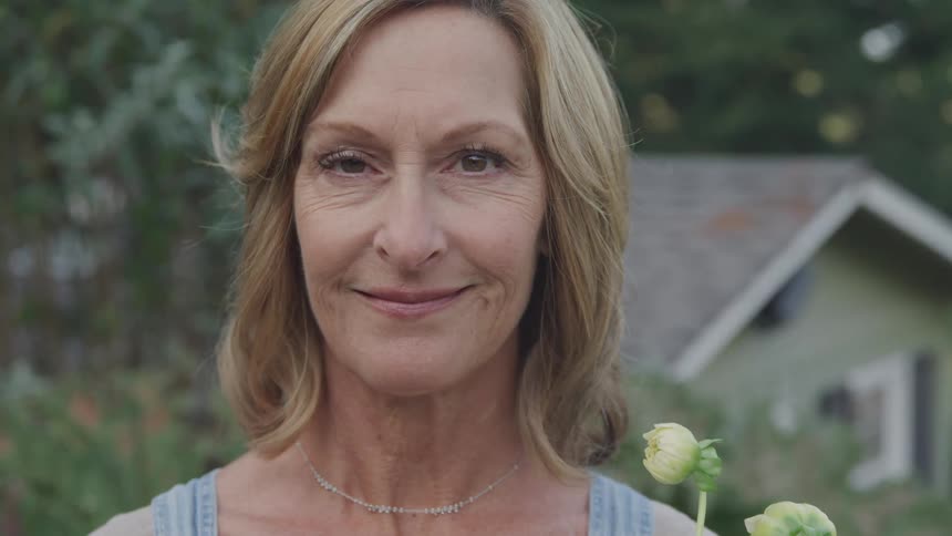 A mature woman with blonde hair a subtle smile and a delicate necklace stands outdoors holding flowers with a building and greenery blurred in the background