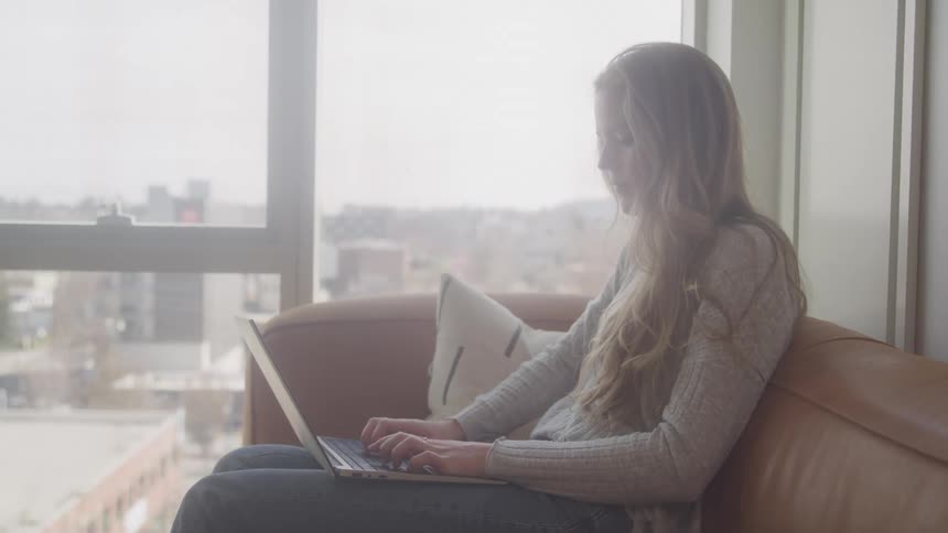 A woman with long blonde hair sits on a couch working on a laptop with a focused expression against a backdrop of a bright window and a cityscape view