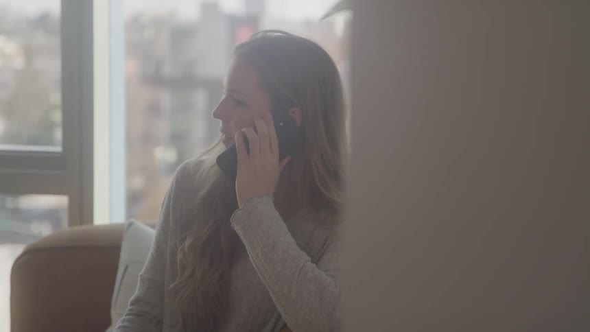 A woman with a gray sweater and long blonde hair talks on a cell phone her profile visible against a blurred background of a window and cityscape