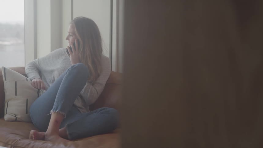A woman with long hair sits barefoot on a couch wearing a light gray sweater and blue jeans talking on a cell phone and looking out of a nearby window