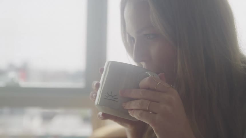 A woman with long blonde hair partially obscured by a soft blur as she holds a mug to her lips with a bright window in the background