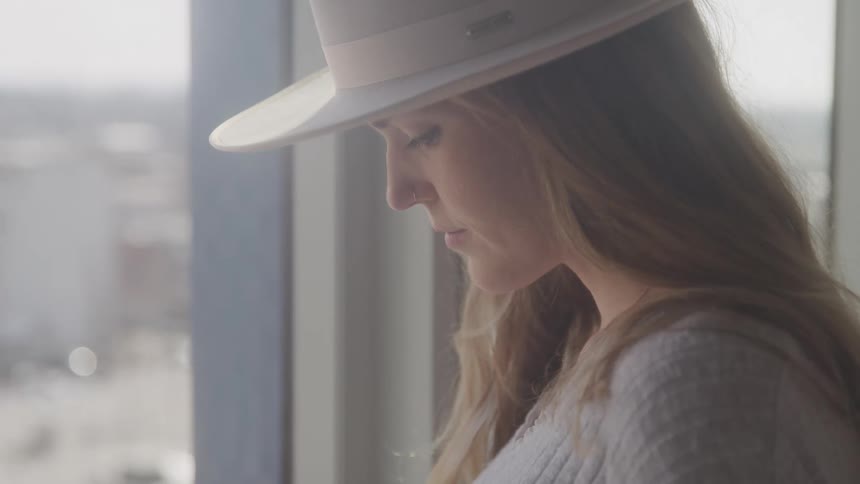 A woman with long blonde hair wearing a white hat looks down with a pensive expression