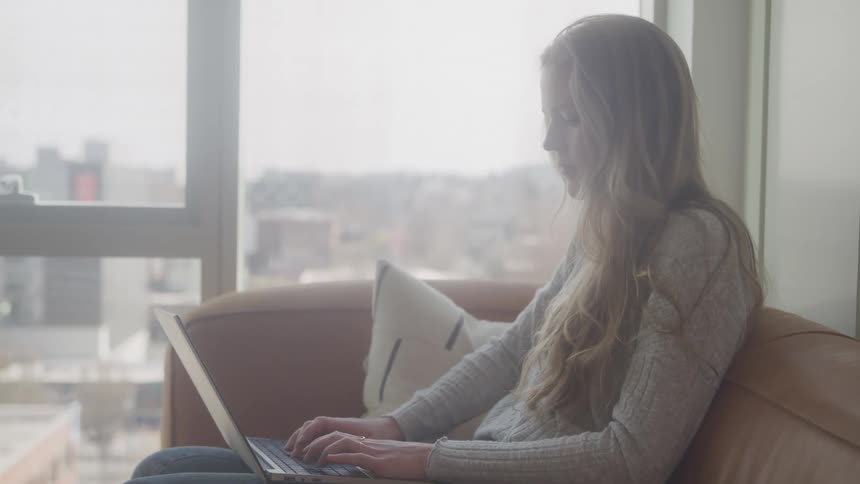 A woman with long blonde hair sits on a couch working on a laptop with a focused expression against a backdrop of a bright window and a cityscape view