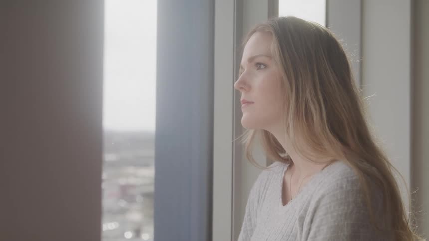 A woman with long lightcolored hair looks out of a window at a cityscape
