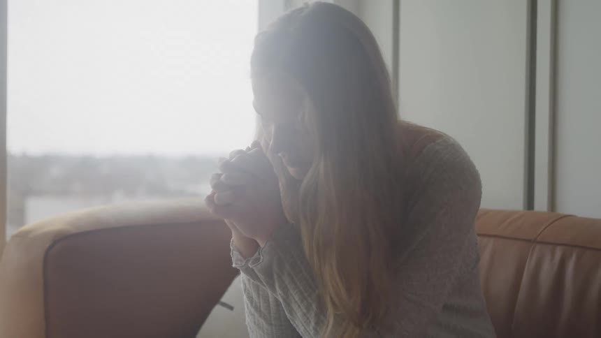 A person with long hair sits on a couch in a brightly lit room hands clasped in prayer lost in thought