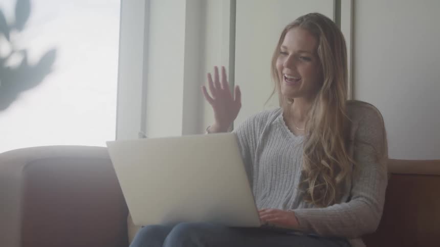 A woman with long blonde hair sits on a couch smiling and waving at a laptop screen having a video call