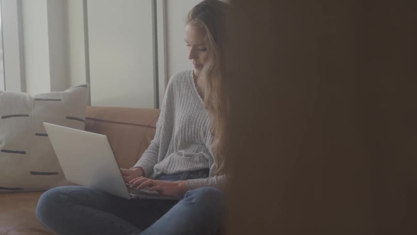 Blonde woman sitting cross legged on her couch types on a laptop during the day