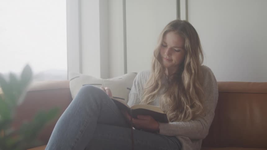 Female sitting on couch near a window skimming through a book