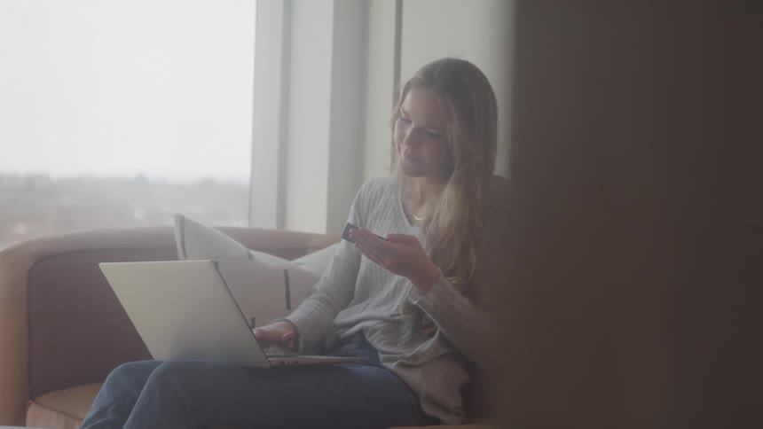 Woman sitting on a couch types on a laptop with one hand and holds a smartphone in the other