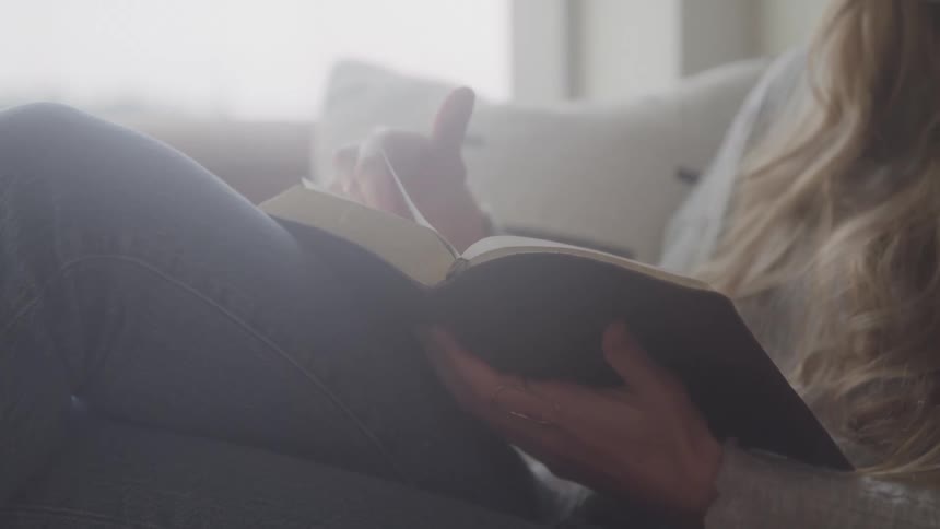 Woman sitting on couch near window reading a hard cover book