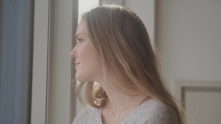 A woman with long blonde hair gazes out of a window her profile illuminated by the soft light
