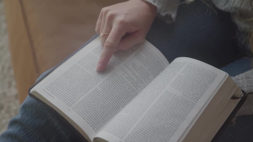 A woman wearing jeans and a light colored sweater points to a passage in an open book she is holding